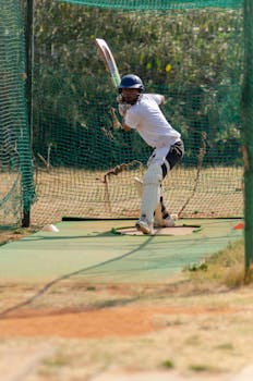 A cricket player practicing a sweep shot in outdoor nets under bright daylight.